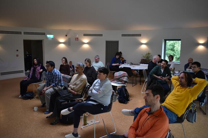 The full group seated in the round room, all facing a screen on a stand.