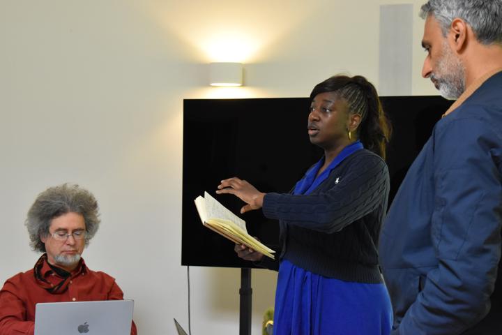 A person standing and speaking from an open notebook, others listening.
