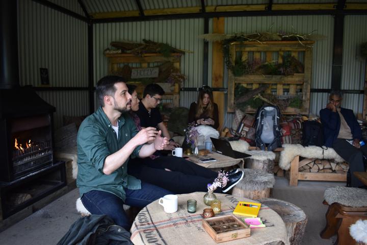 A group seated around a wood stove in the shed, mugs in hand.