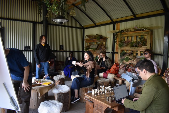 A session in the shed: one person standing, others on sheepskin-covered log stools, one speaking with a mug in hand and another at a laptop.