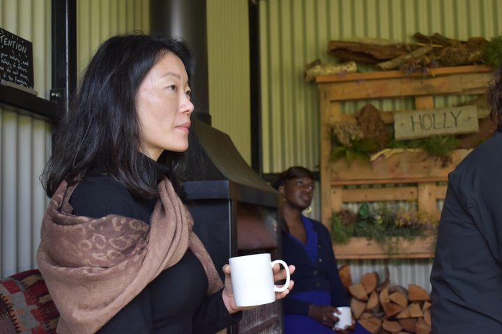 A person in a patterned scarf listening intently, mug in hand, beside the shed's stove.