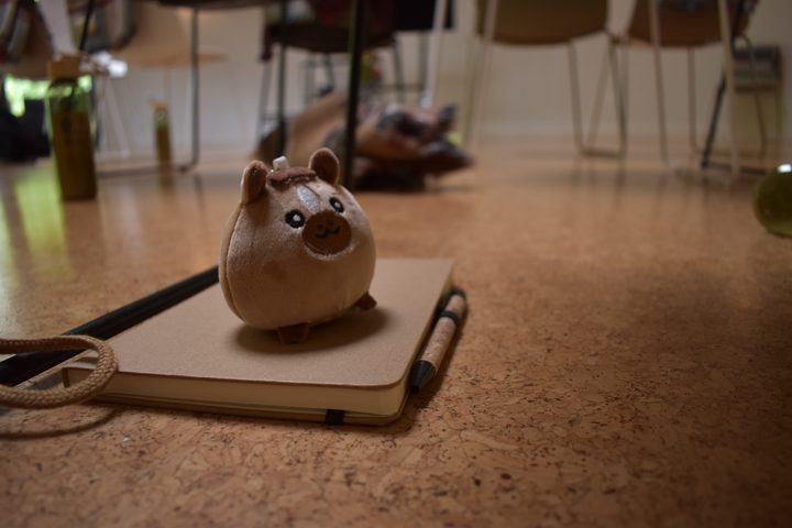 Close-up of the small plush mascot sitting on a notebook with a pencil, chairs blurred behind.