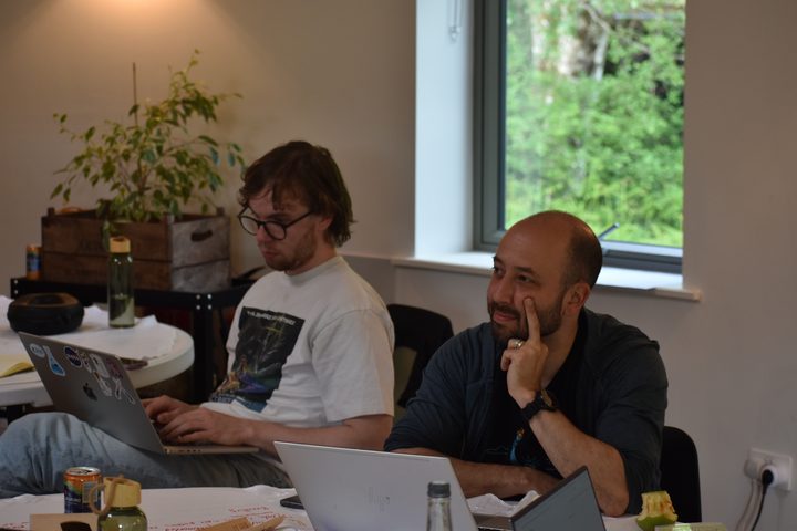 Two people at a table, one typing on a laptop, the other listening with a hand at their cheek, green trees in the window behind.