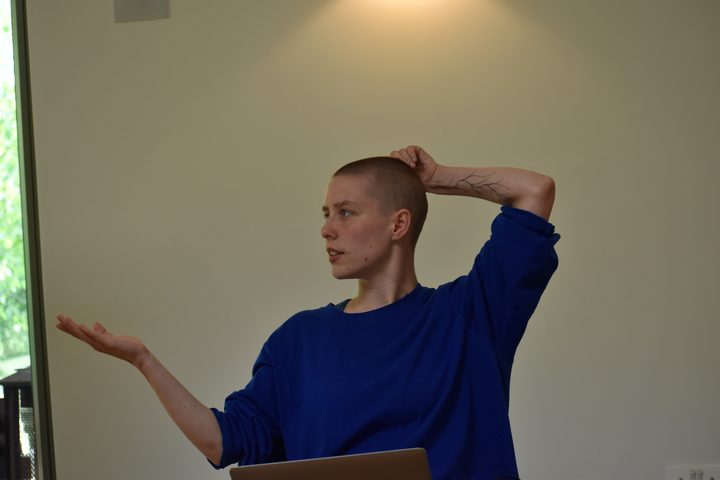 A person in a blue top speaking, one hand raised and the other resting on their head.
