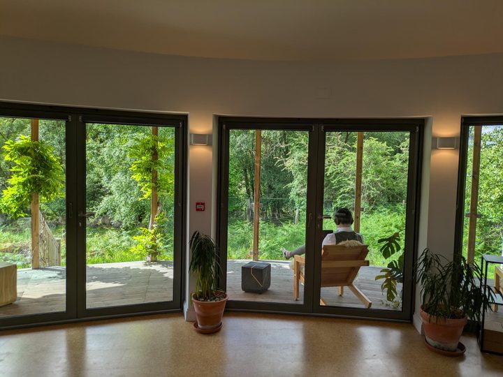 The round room's glass doors open onto a wooden deck, where one person sits in the sun looking out at the trees.